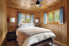 A wooden-paneled bedroom with a bed, two bedside lamps, a ceiling fan, and windows with curtains.