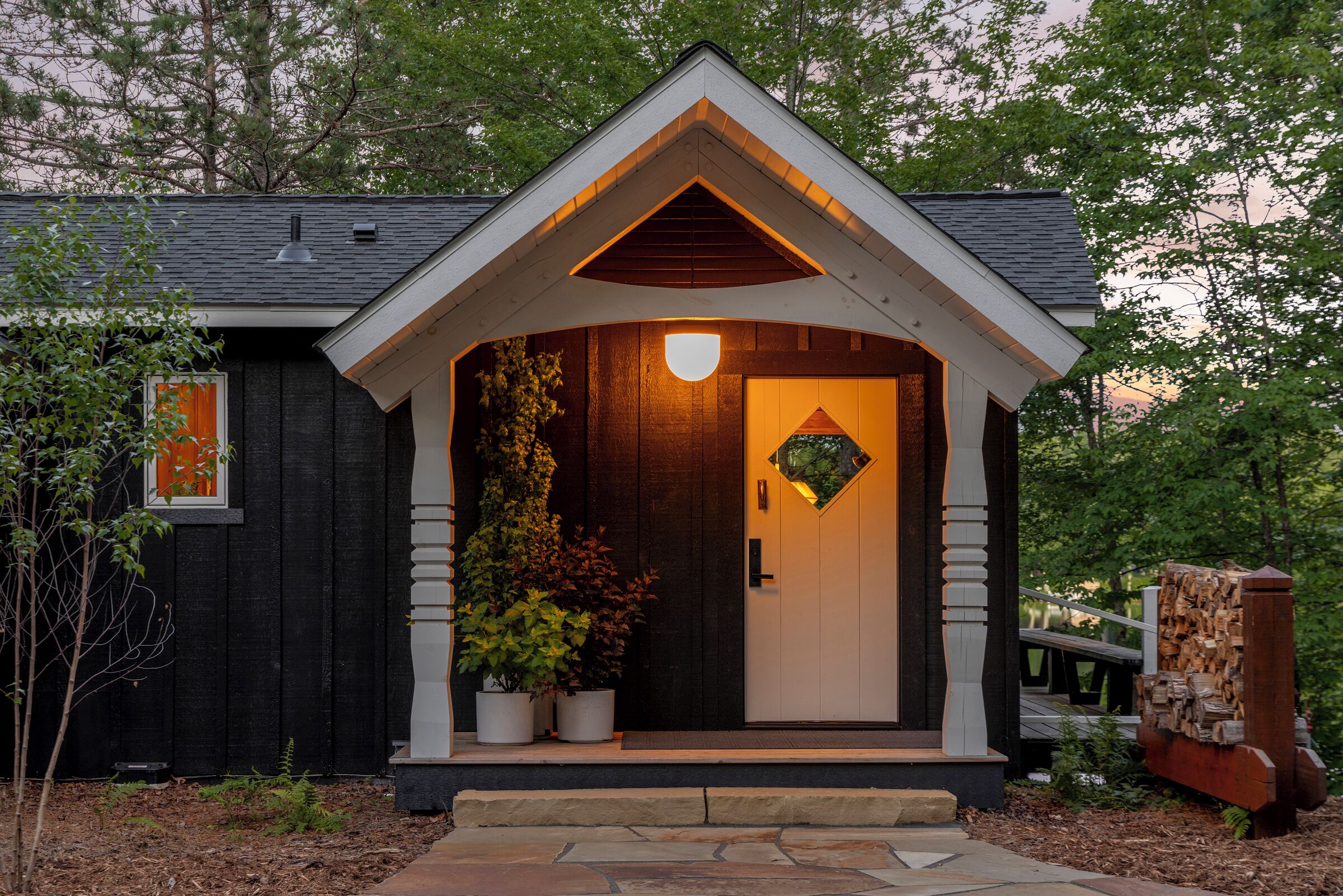 A small, dark-colored building with a white door and a triangular roof. There are potted plants on the porch and a wooden bench to the right.