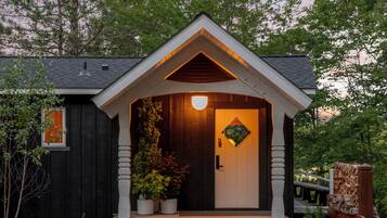 A small, dark-colored building with a white door and a triangular roof. There are potted plants on the porch and a wooden bench to the right.
