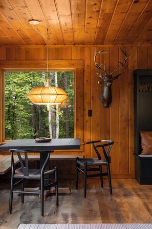 A wooden interior with a dining table, two chairs, a hanging light fixture, and a mounted deer head.