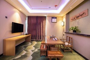 Habitación de hotel con una larga mesa de madera para comer, sillas de madera, un televisor de pantalla plana montado en la pared, un banco y un ventanal con cortinas.