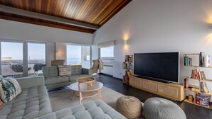 A modern living room with a large flat-screen TV, comfortable seating, a bookshelf, and a view of the sea through large windows.