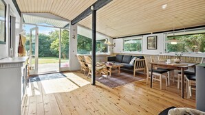A spacious living area with a wooden floor, a sofa, a dining table, and chairs. There is a large window providing a view of the outdoors. - 4 bedroom cozy home in Jægerspris (Jægerspris)