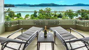 A balcony with two lounge chairs, a table, and a bottle of champagne. The balcony overlooks a body of water with trees and hills in the background.