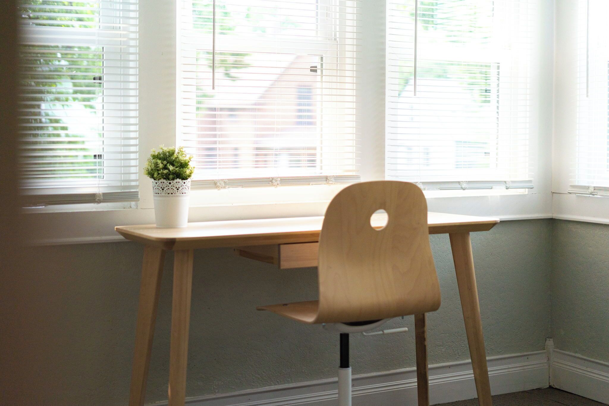 A corner desk with a wooden chair, a small potted plant, and a window with blinds.