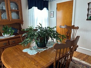 Una mesa de comedor de madera con un mantel de encaje, rodeada de sillas de madera. Hay una planta en maceta sobre la mesa.