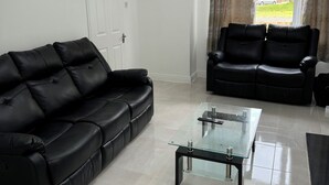 A modern living room with a black leather sofa, a glass coffee table, and a view of a residential street through a window with curtains. - 4-bedroom house in charming Lucan with WiFi (Lucan)