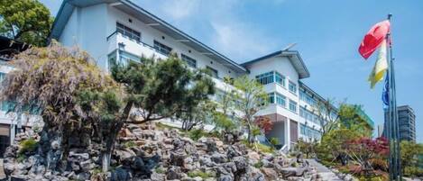 Ein modernes Hotel mit heller Fassade, einem Felsenbeet und einer Fahnenstange mit Flaggen.