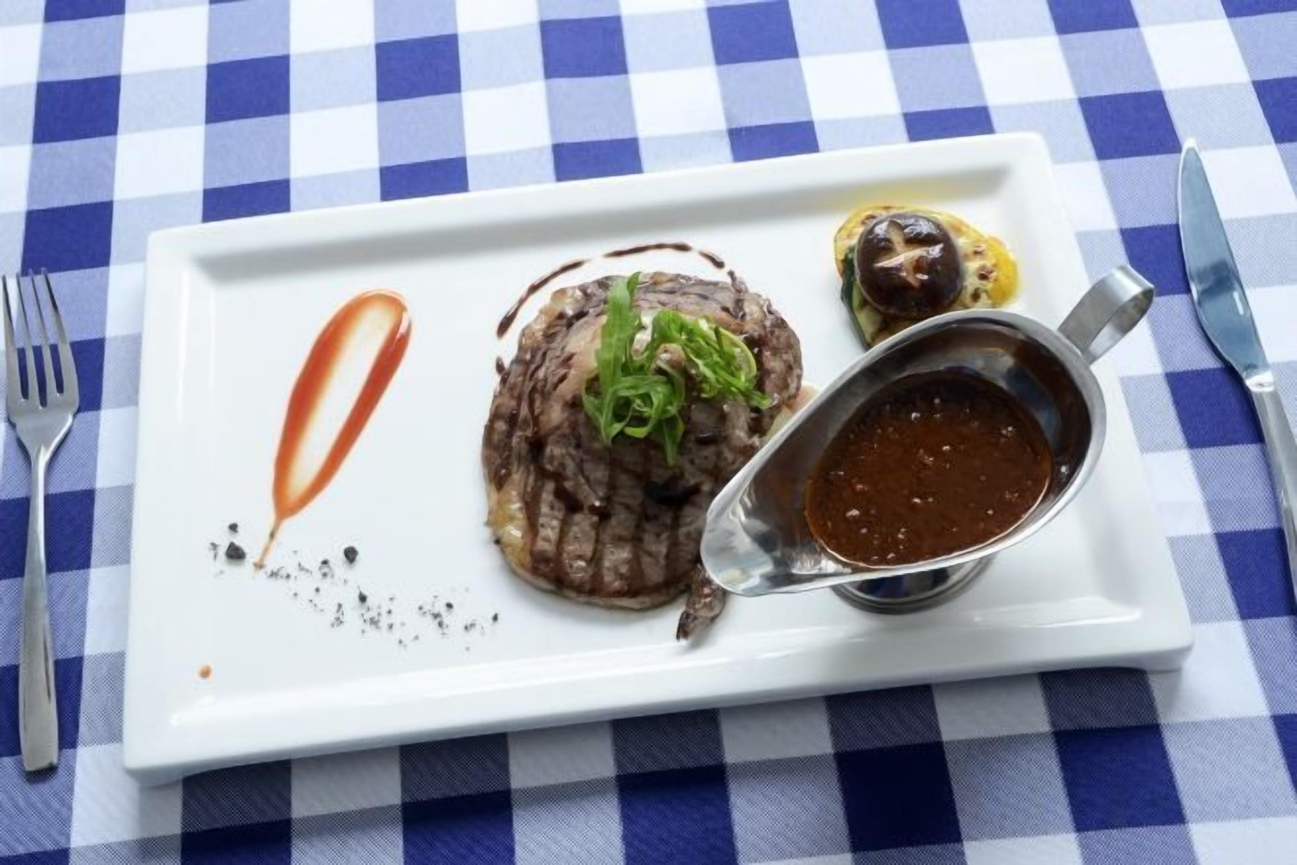 Un piatto bianco rettangolare con una bistecca, una salsa e una piccola ciotola di salsa su una tovaglia a quadretti blu e bianchi.