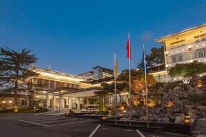 A modern hotel with a glass facade, a landscaped entrance, and multiple flags. - Riverside Garden Holiday Hotel (Hangzhou)