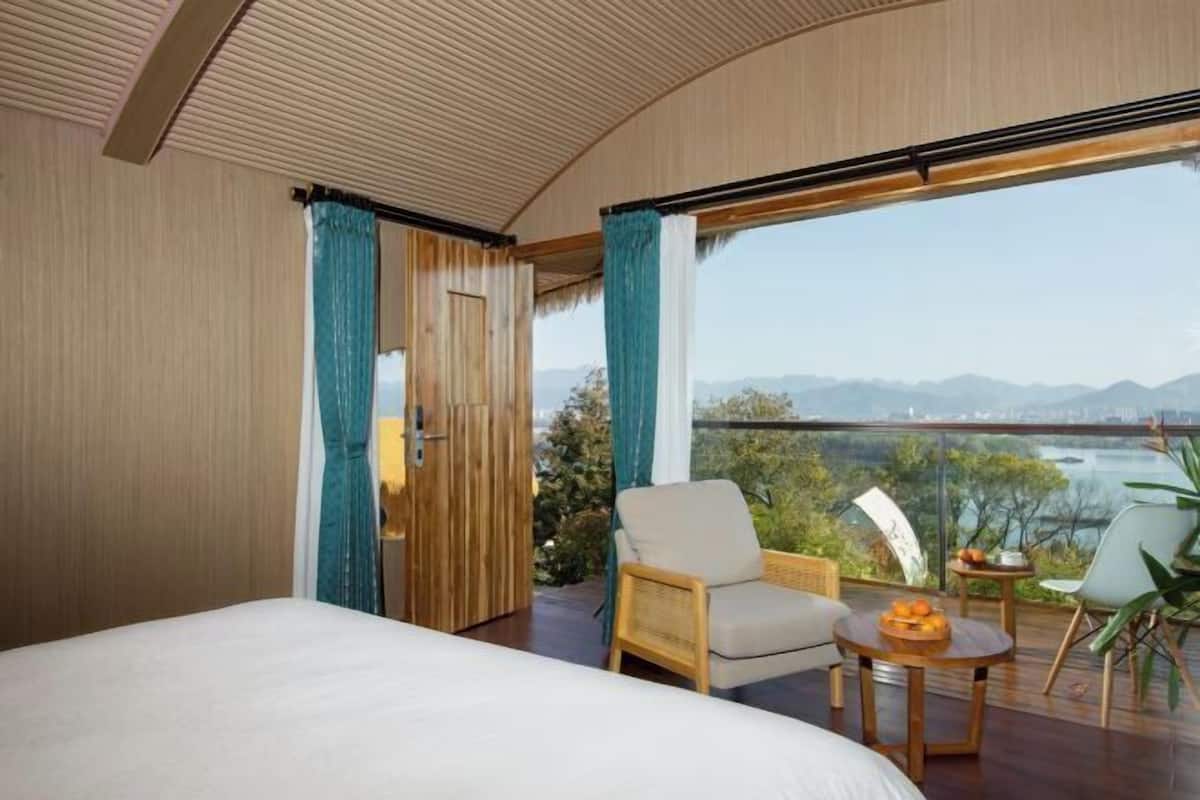 Un dormitorio con una cama grande, una silla, una mesita con fruta y vistas a un cuerpo de agua y montañas.