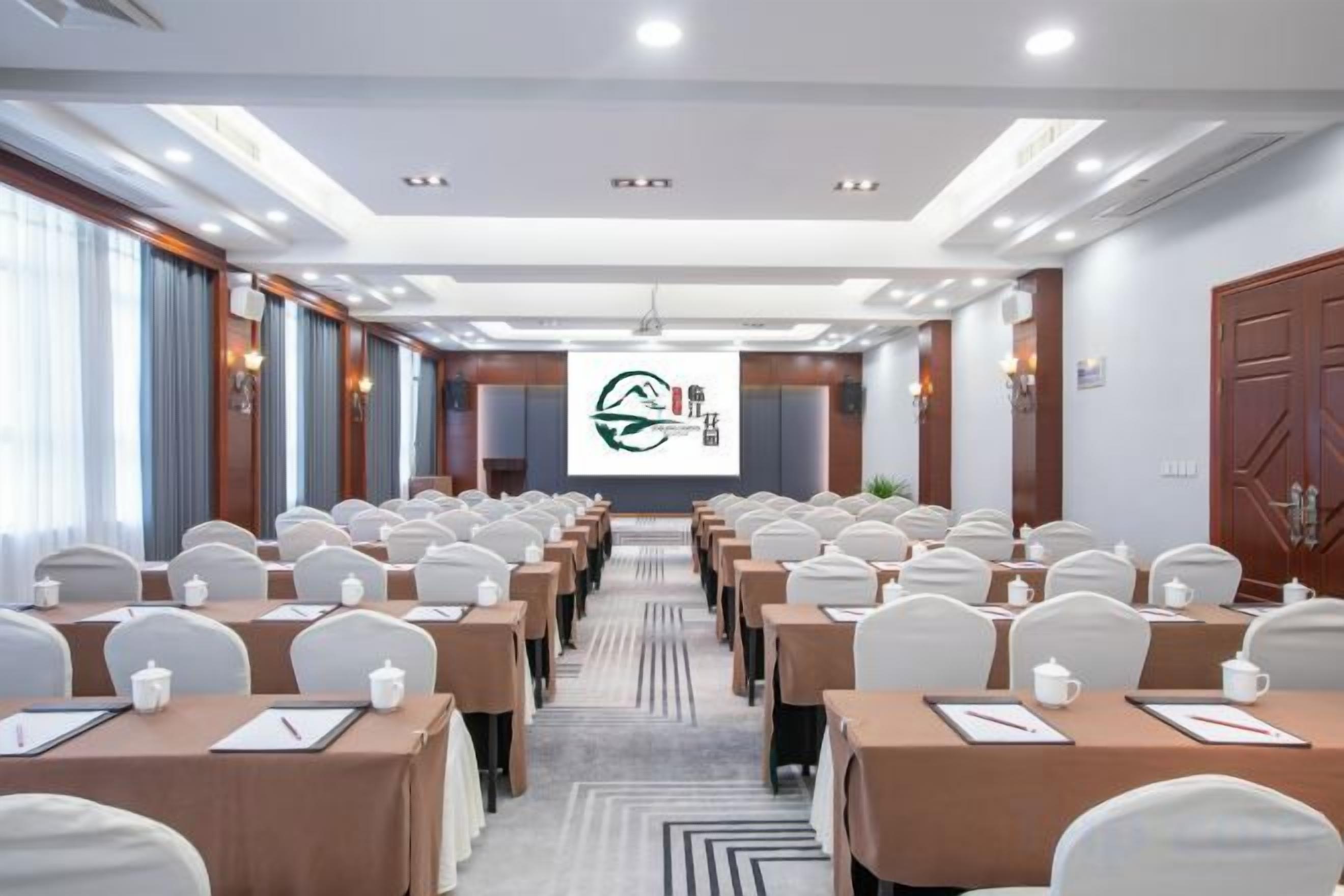 A conference room with rows of tables and chairs, a projector screen, and a presentation on the screen.