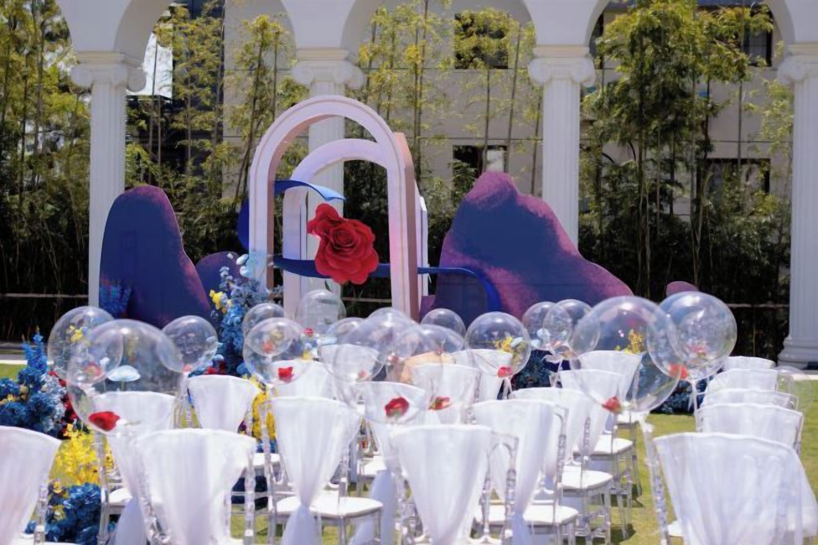 Ambiente externo com cadeiras brancas dispostas em fileiras, um arco decorativo com uma rosa vermelha e um cenário de árvores e um edifício.