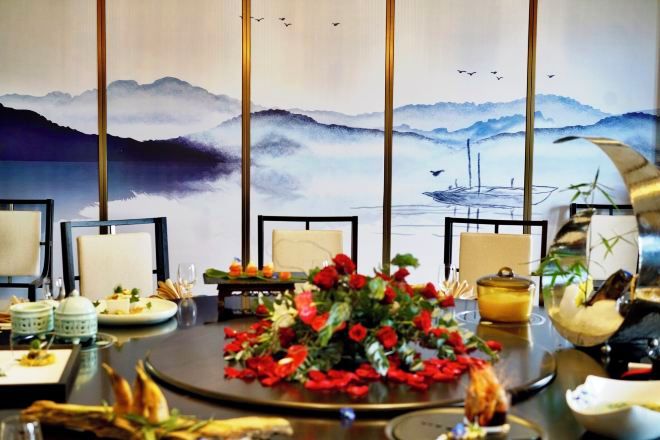 A dining table with chairs, a floral centerpiece, and a painting of mountains and water in the background.