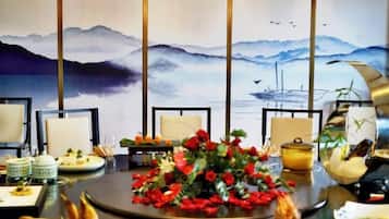 A dining table with chairs, a floral centerpiece, and a painting of mountains and water in the background.