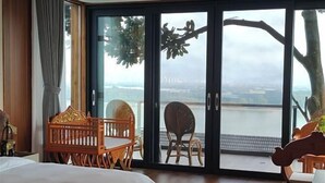 Habitación de hotel con un gran ventanal corredero que da a un balcón con vistas a una masa de agua y un árbol. Se observa una cama con ropa blanca y una silla de madera con cojín.