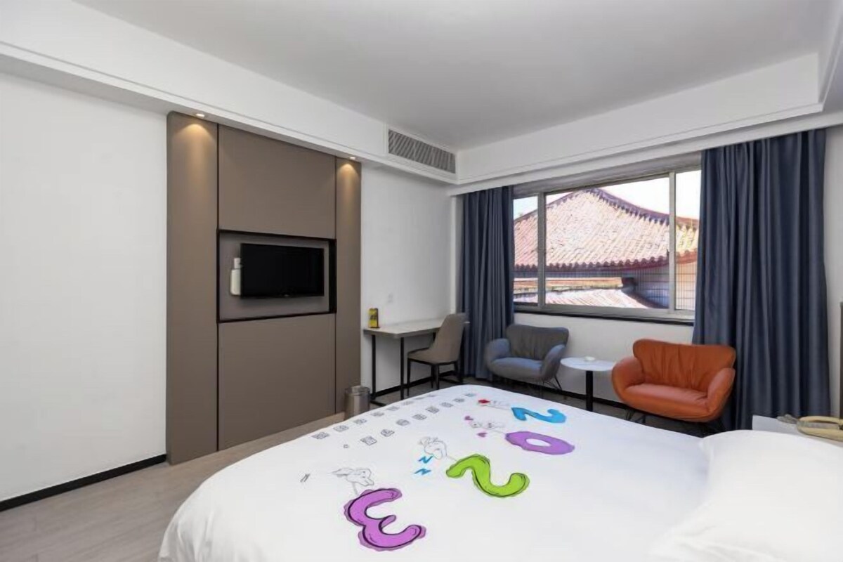 A modern hotel room with a bed, a TV, a desk, and a view of traditional architecture.