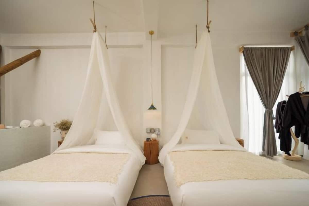 Ein Zimmer mit zwei Betten, einem Holzkopfstück, einem kleinen Tisch mit Lampe und einem Fenster mit Vorhängen.