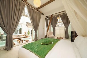 Une chambre dotée d’un grand lit, d’un tapis vert, d’un fauteuil et offrant une vue sur un balcon aménagé avec du mobilier d’extérieur.