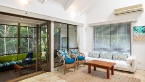 Een moderne woonkamer met een glazen deur die leidt naar een balkon, een witte bank, twee rieten fauteuils met blauwe kussens, een houten salontafel en een groot raam met zonwering.