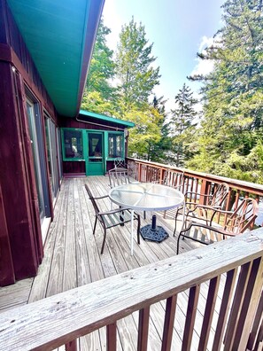 A wooden deck with a glass table and chairs, a covered area with a green roof, and a view of trees and a body of water.