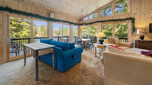 A cozy cabin interior with a wooden ceiling, large windows, a dining area, and a seating area with a blue sofa and beige armchair. - Lakefront Escape, On Snowmobile Trail, Near Town (Old Forge)