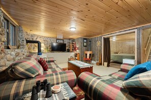 A cozy cabin interior with a stone wall, wooden ceiling, plaid sofas, a television, and a fireplace.
