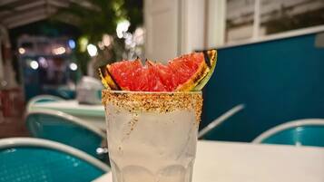 Un vaso alto con borde de sal, que contiene un líquido transparente y está decorado con una rodaja de fruta, sobre una mesa blanca en una habitación con paredes azules y una piscina al fondo.
