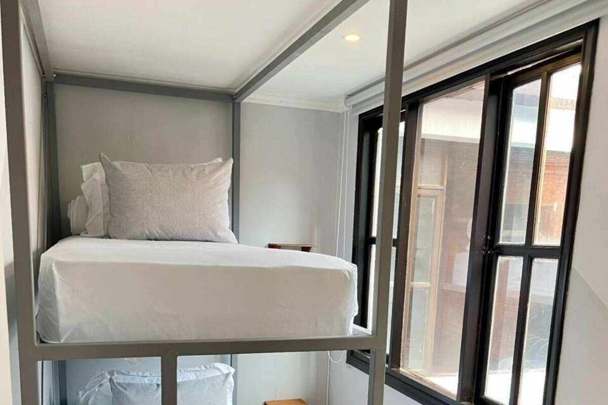 A bunk bed with a ladder, a window with blinds, and a small wooden stool.
