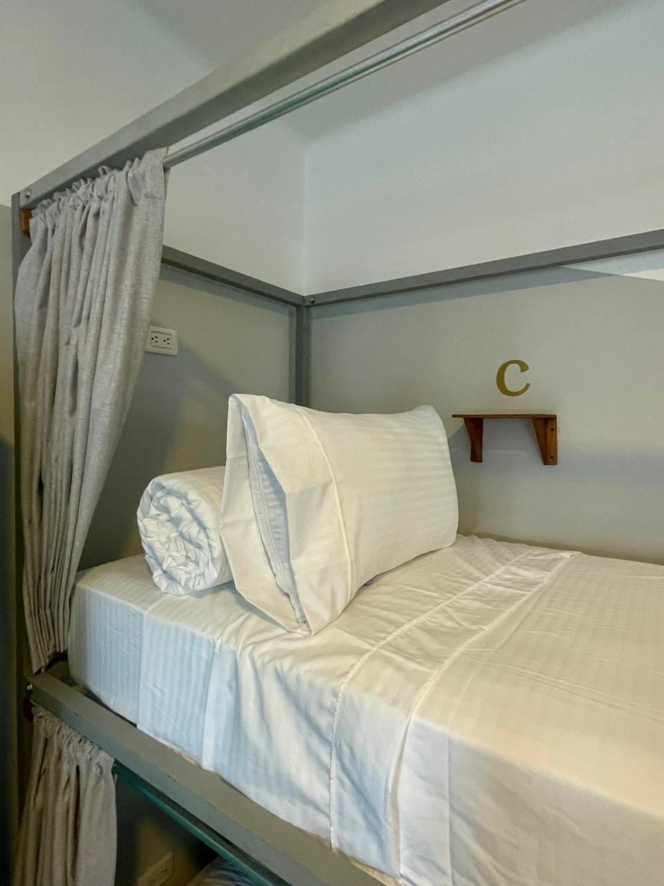 Una cama litera con almohada y manta, una cortina y un pequeño estante con la letra 'C'.