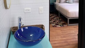 Une salle de bain moderne avec un lavabo bleu, un mur en briques blanches et un lit visible à travers une porte ouverte.