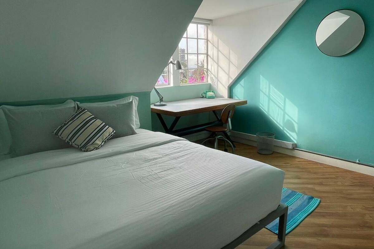 A bedroom with a bed, a desk, a chair, a window, and a mirror.
