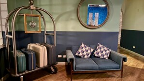 Une pièce avec un canapé bleu, deux coussins à motifs, un chariot à bagages avec des valises, un miroir rond et un mur orné d’un motif bleu.
