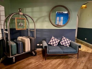 Una habitación con un sofá azul, dos almohadas estampadas, un carro de equipaje con maletas, un espejo redondo y una pared con un diseño azulado.