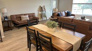 A living room with a brown leather sofa, a dining table with chairs, a small side table with a lamp, and a window with a view of the outside. - Brand New Luxury Townhouse Getaway in Lewiston, Idaho!   (Lewiston)