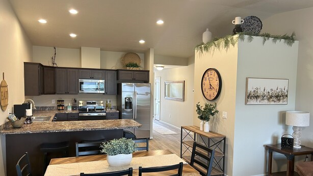 A modern kitchen with dark cabinets, stainless steel appliances, and a granite countertop. There is a dining area with a wooden table and chairs, and a small side table with a lamp. - Brand New Luxury Townhouse Getaway in Lewiston, Idaho!   (Lewiston)
