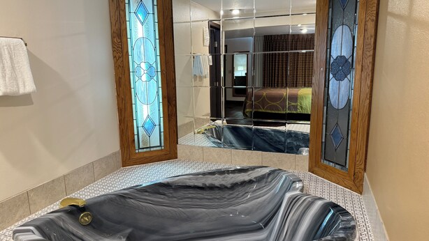 Ein Badezimmer mit einer großen, ovalen Badewanne aus schwarzem Marmor, einer Glastür mit Holzrahmen und einem Spiegel, der ein Schlafzimmer mit Bett und Vorhängen reflektiert.
