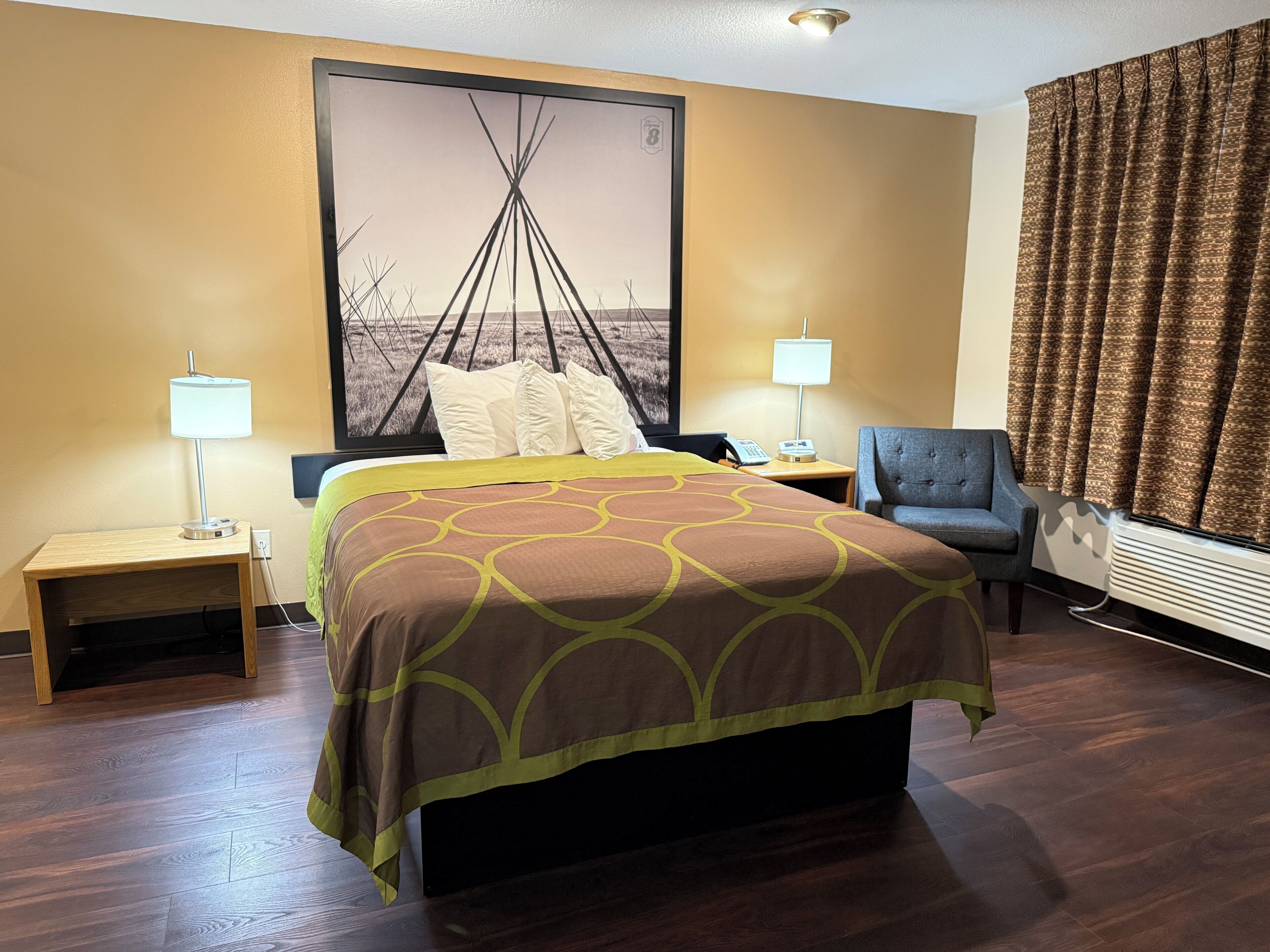 A hotel room with a large bed, two bedside tables with lamps, a chair, a large window with curtains, and a wall-mounted air conditioner.