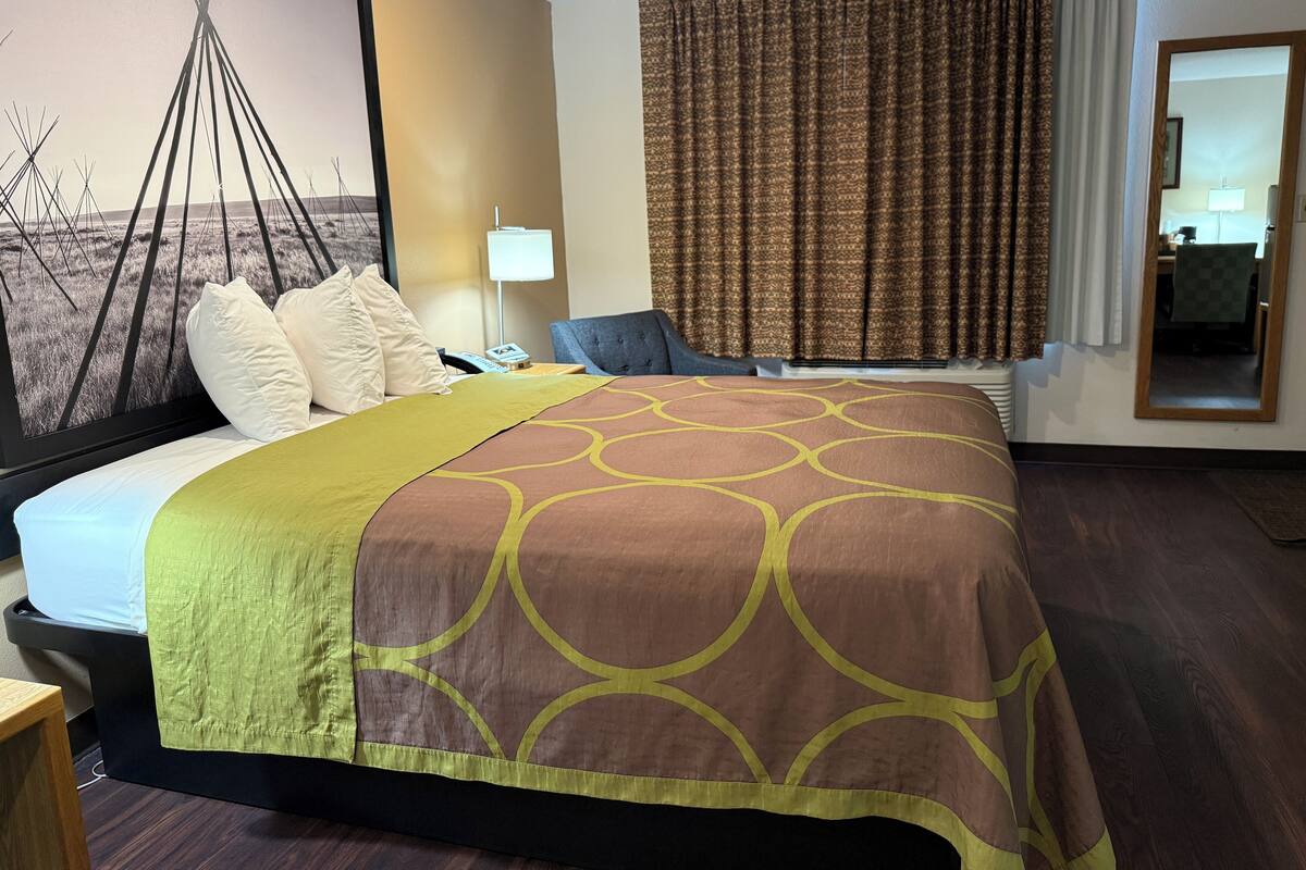 Une chambre d’hôtel avec un lit, une table de chevet, une lampe, une chaise et une grande fenêtre avec des rideaux.