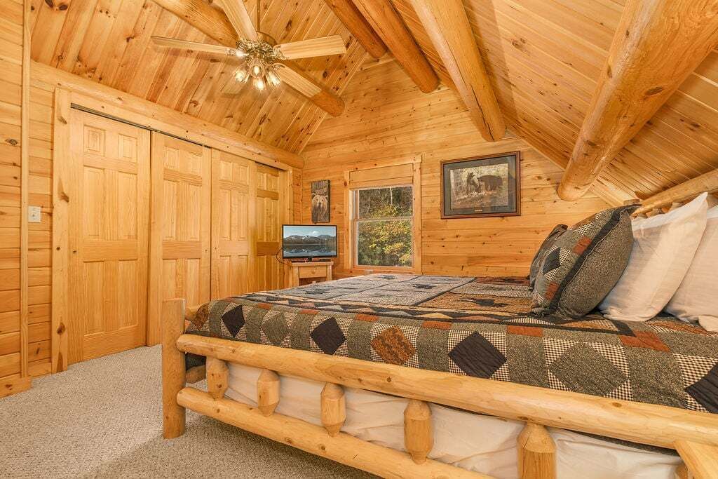 Un dormitorio tipo cabaña de madera con una cama de madera, un ventilador de techo, un televisor y un cuadro enmarcado en la pared.