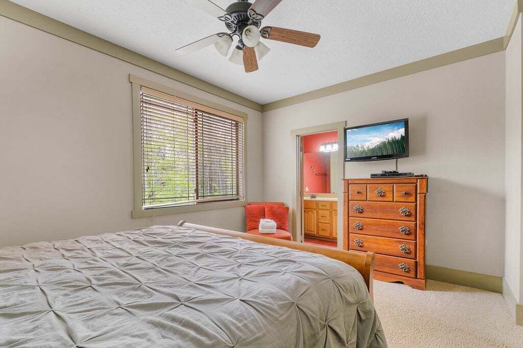 A bedroom with a bed, a ceiling fan, a dresser, a television, and a window with blinds.