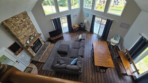 A spacious living room with a stone fireplace, large windows, and a wooden dining table. - Family Mountain Escape w/ Hot Tub & Game Room (Oakland)