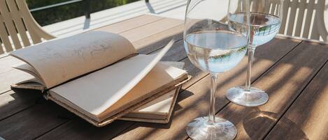 Ein Holztisch mit einem offenen Buch und zwei Gläsern Wasser, platziert auf einer Terrasse mit Meerblick.