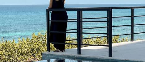 A woman in a black dress standing by a pool with a view of the ocean.