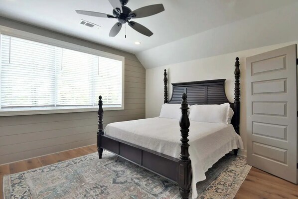 A bedroom with a four-poster bed, a ceiling fan, a large window with blinds, and a patterned rug. - Boots & suits lakehouse getaway
(Cordele)