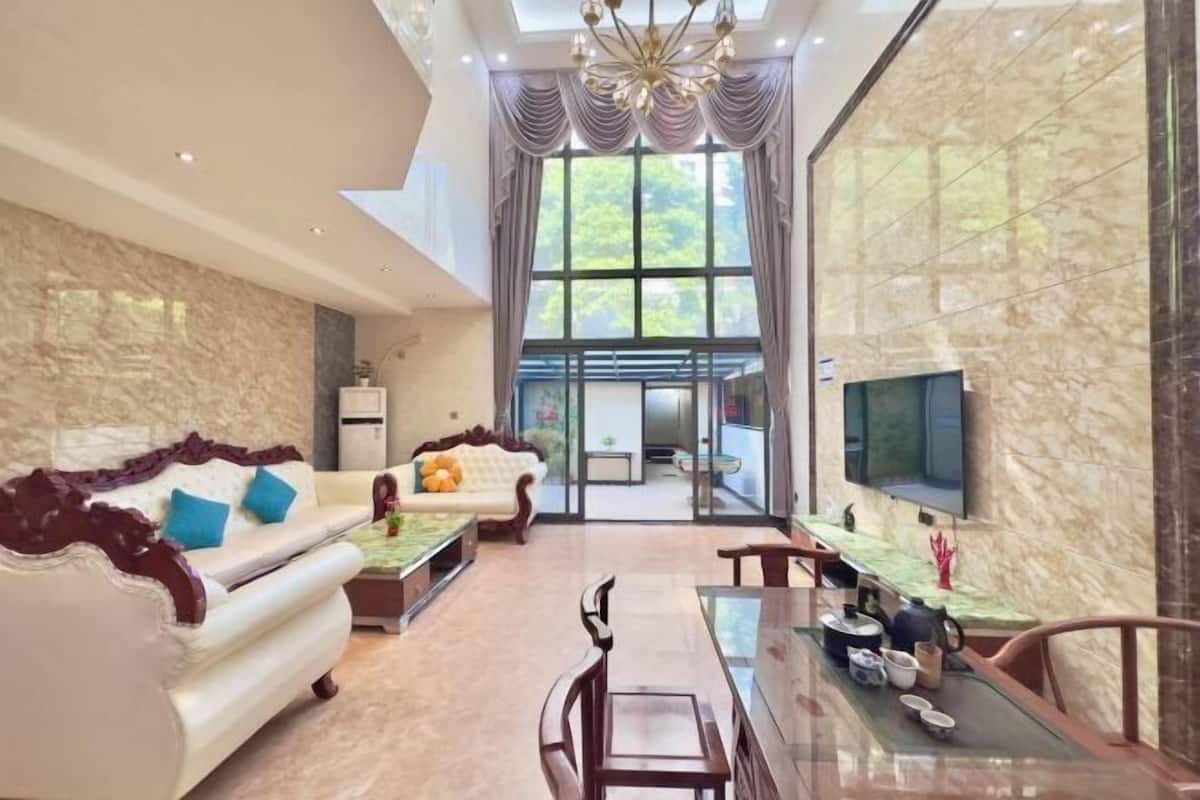 A spacious living room with a high ceiling, a large window, a sofa, a dining table, and a TV.