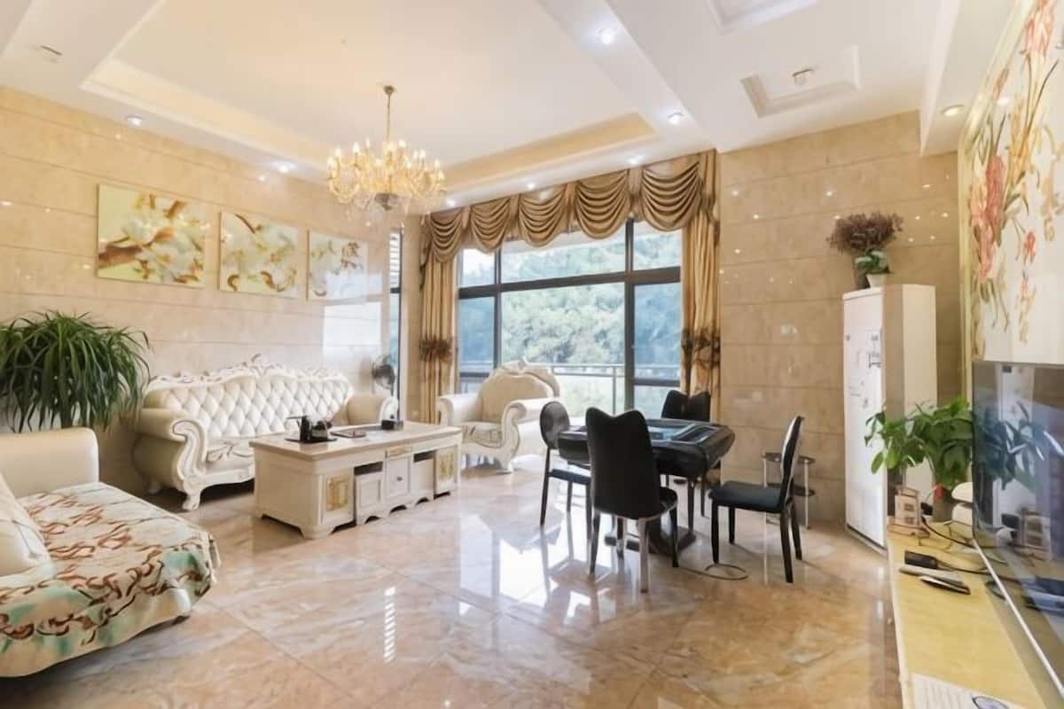 A spacious living room with a large sofa, dining table, and chairs, a chandelier, and large windows.