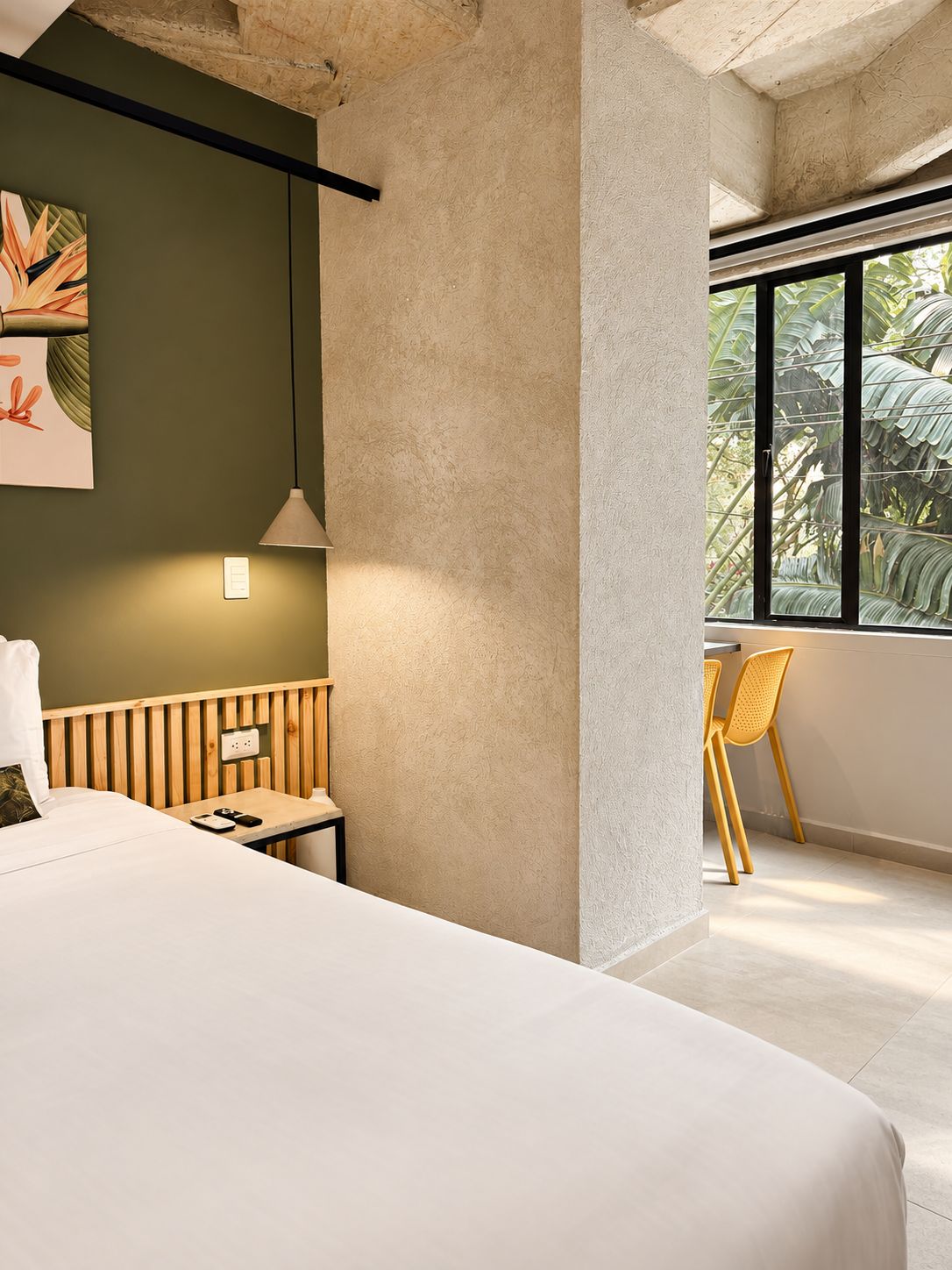 A modern hotel room with a large bed, a desk with a lamp, a chair, and a view of greenery through a window.