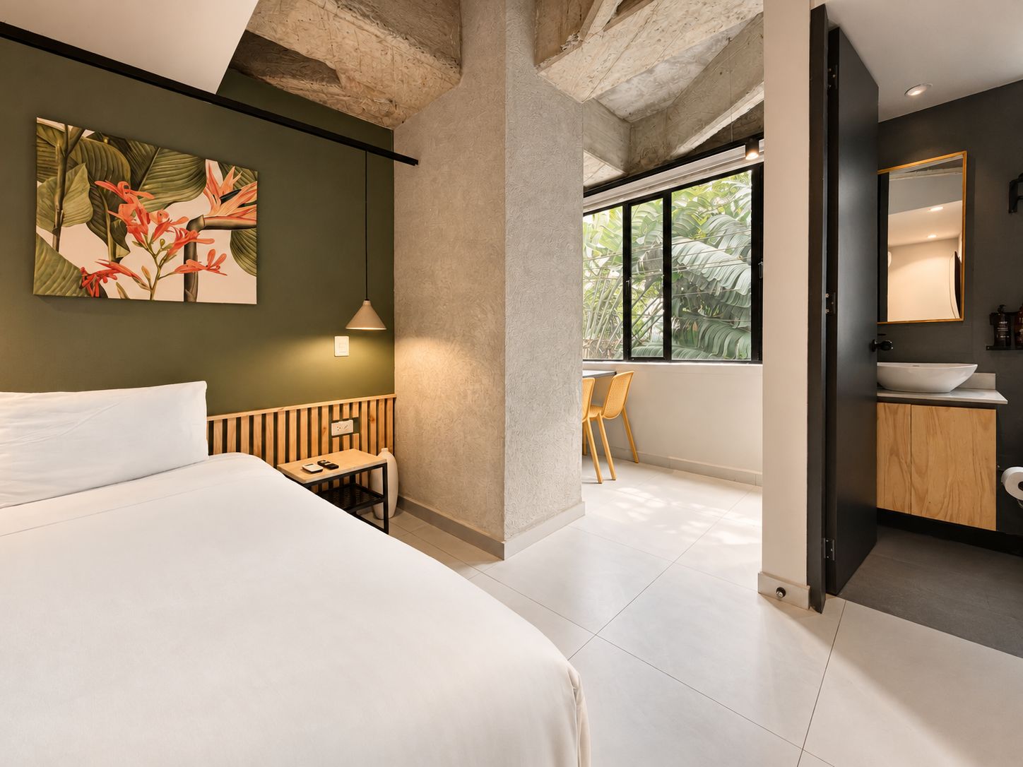 A modern hotel room with a large bed, a desk, a chair, a window with a view of greenery, and a bathroom with a sink and mirror.