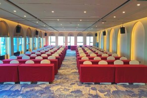 Un salón de conferencias con filas de mesas y sillas, ventanas amplias y una alfombra estampada.